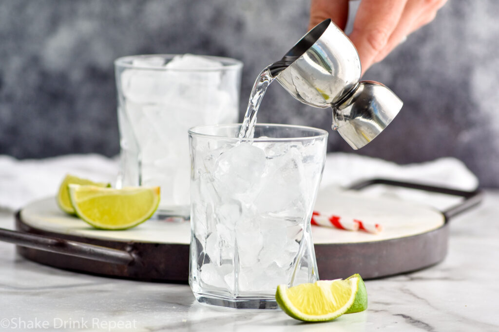 man's hand pouring jigger of rum into a glass of ice to make a Cuba Libre surrounded by slices of lime