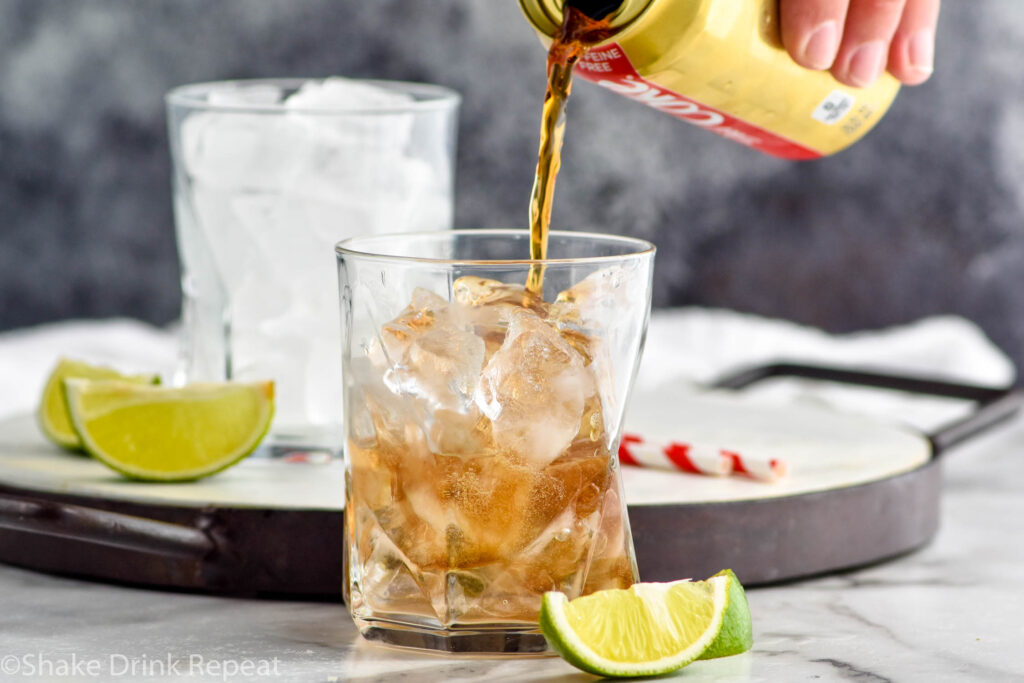 man pouring can of coke into a glass of ice and rum to make a Cuba Libre cocktail surrounded by straws and slices of lime
