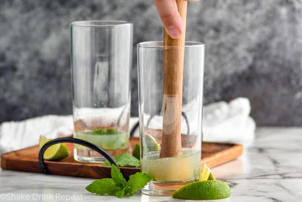 Man's hand using a wooden muddler to muddle mint in simple syrup and lime juice in a glass to make a Mojito recipe. Surrounded by fresh mint leaves and slices of lime