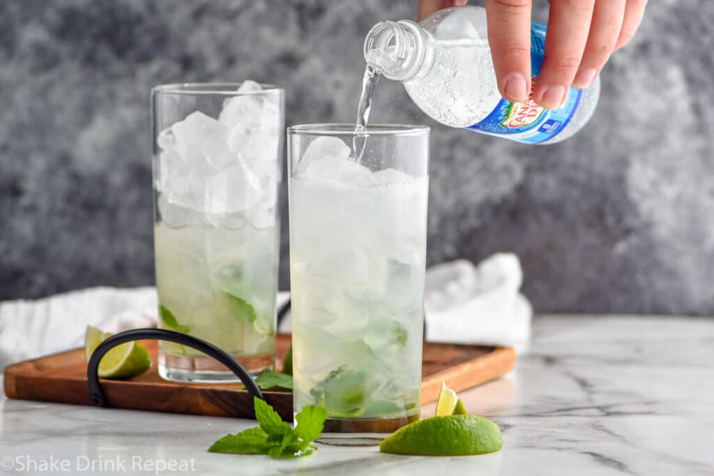 man pouring bottle of club soda into a glass of ice and Mojito ingredients surrounded by fresh mint leaves and lime wedges