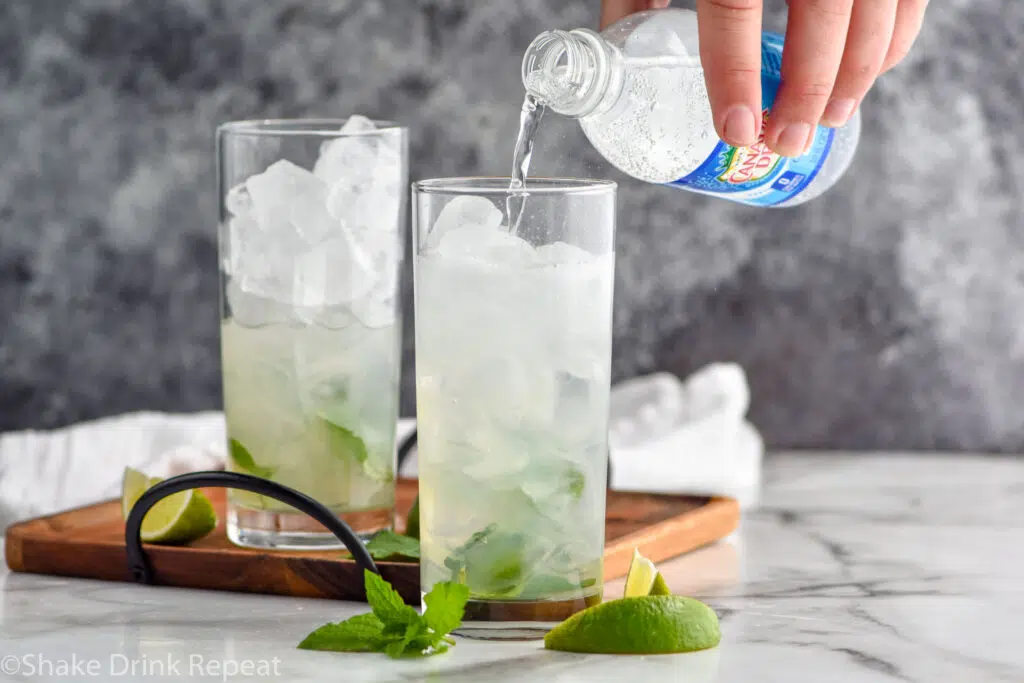 man pouring bottle of club soda into a glass of ice and Mojito ingredients surrounded by fresh mint leaves and lime wedges