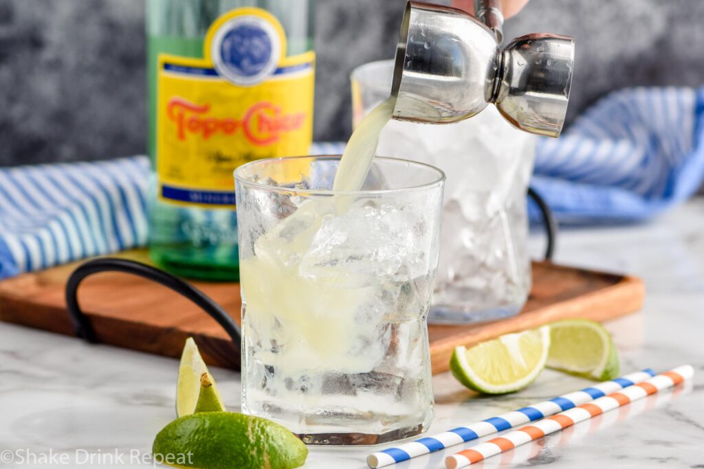 Jigger of lime juice being added to glass of Ranch Water ingredients and ice surrounded by fresh lime wedges, straws, and bottle of Topo Chico