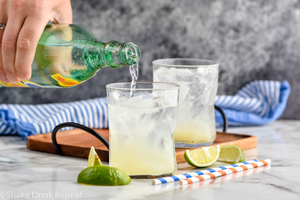 man pouring bottle of Topo Chico into a glass of Ranch Water ingredients and ice surrounded by fresh lime wedges and straws