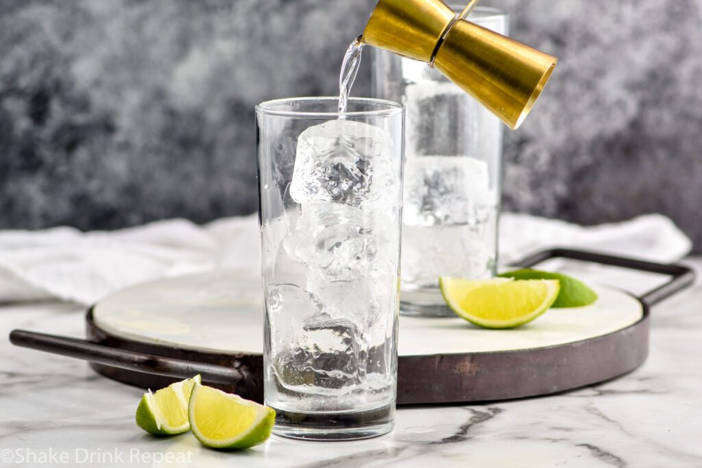 jigger of rum pouring into a glass of ice to make a rum and coke recipe surrounded by slices of lime