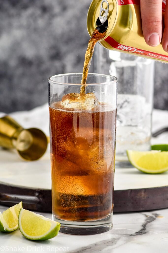 man's hand pouring can of coke into a glass of ice and rum surrounded by slices of lime and gold jigger