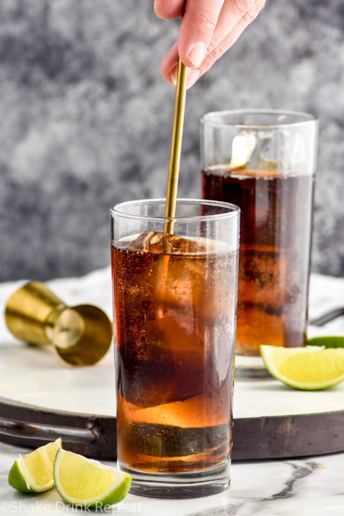 man's hand stirring glass of rum and coke with ice surrounded by slices of lime and a gold jigger