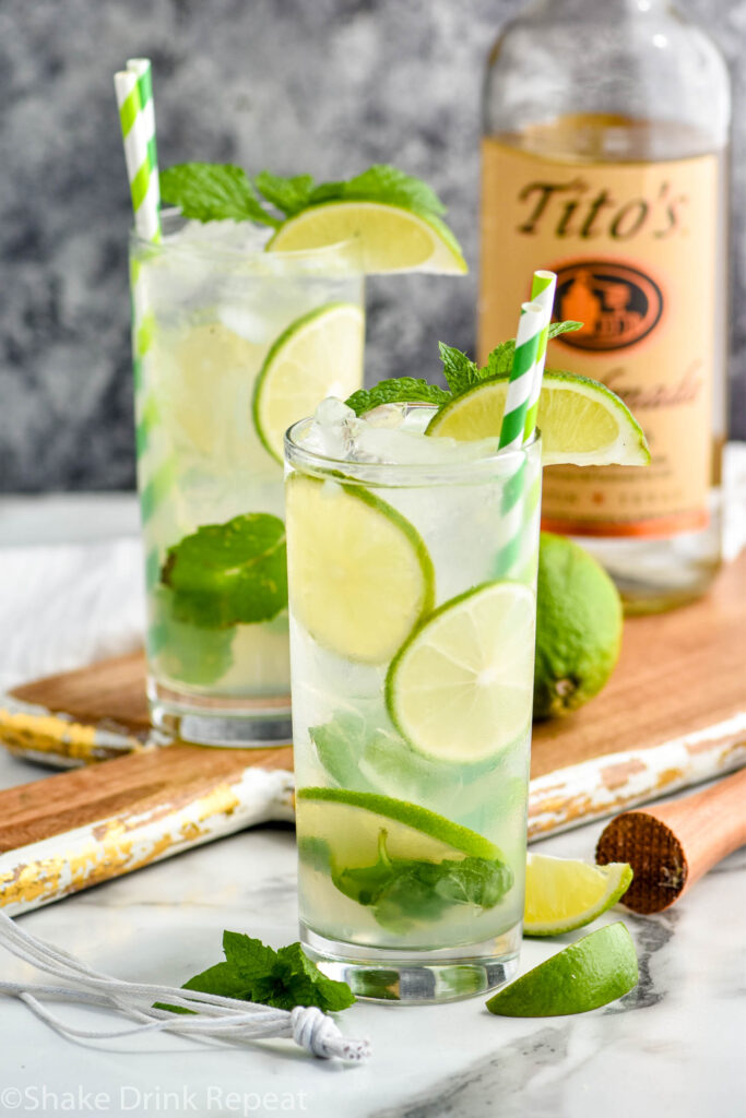 two glasses of vodka Mojito with ice, straws, slices of lime, and fresh mint leaves with bottle of Tito's vodka and a cocktail muddler in the background