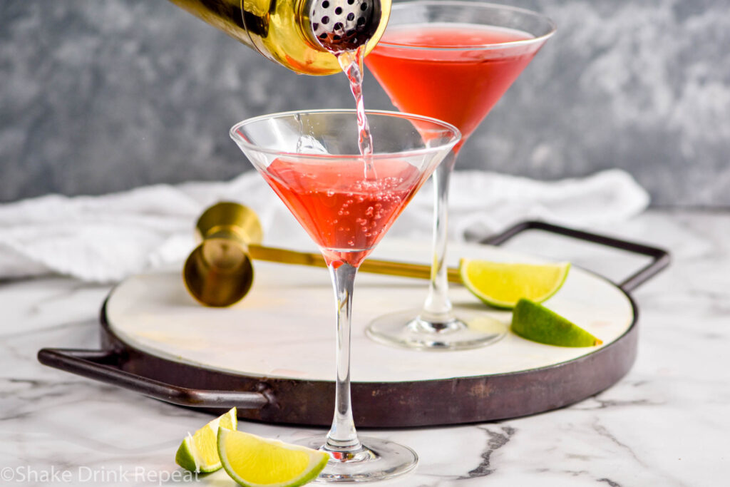 shaker of Woo Woo ingredients pouring into martini glass surrounded by slices of lime and jigger