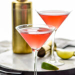 two martini glasses of Woo Woo cocktail with slices of lime, jigger, and cocktail shaker in the background