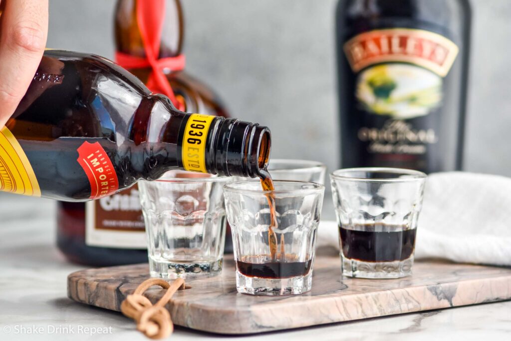 man's hand pouring bottle of Kahlua into a shot glass to create a B-52 cocktail recipe with bottles of Baileys and Grand Marnier in the background