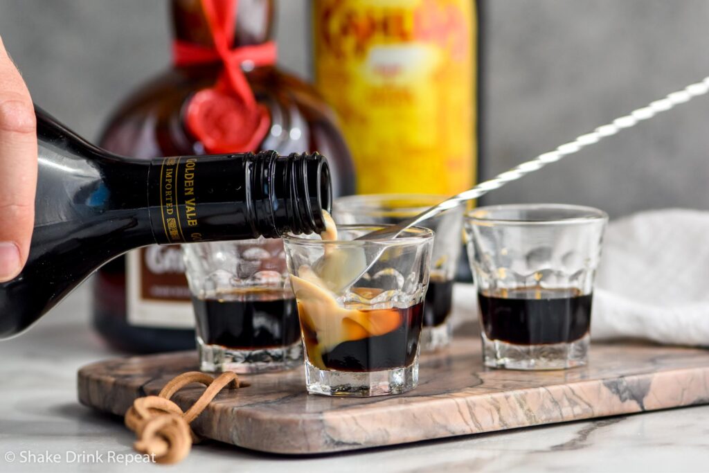 man's hand pouring bottle of Bailey's over a spoon into a shot glass of B-52 ingredients with bottles of Grand Marnier and Kahlua in the background