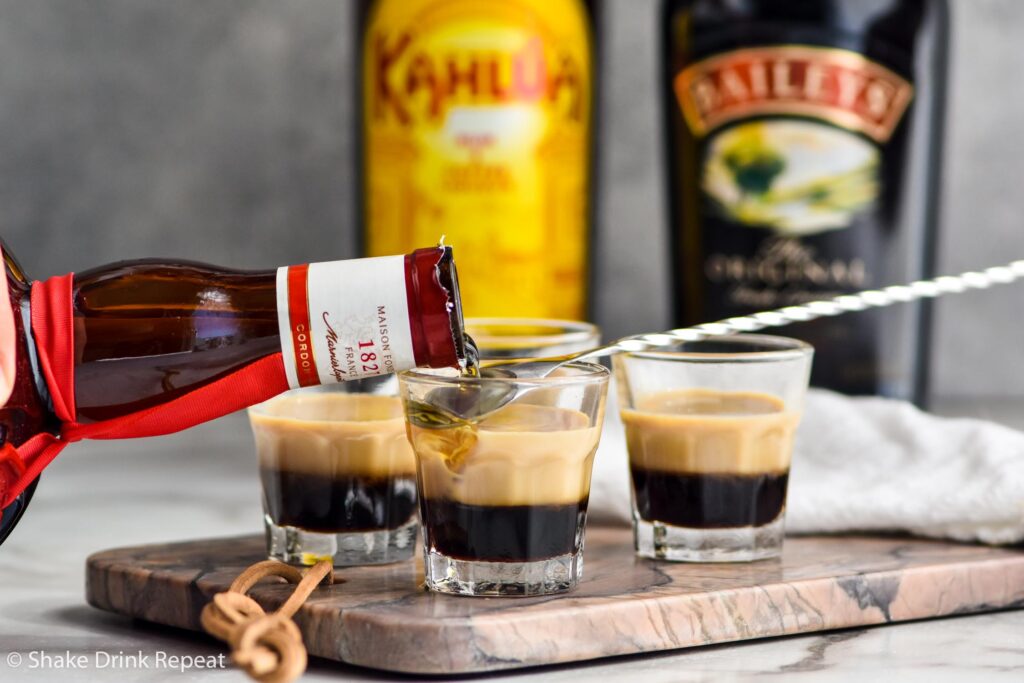 pouring bottle of Grand Marnier over a spoon into a shot glass of B-52 ingredients with bottles of Kahlua and Baileys in the background