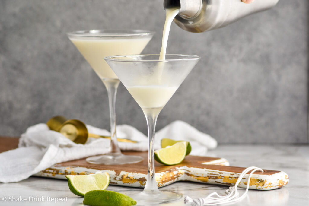 man's hand pouring shaker of Key Lime Martini ingredients into a martini glass surrounded by slices of lime and jigger