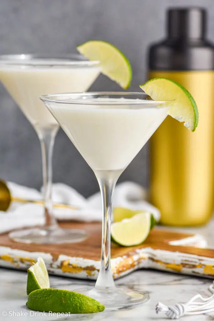 two martini glasses of Key Lime Martini garnished with lime wedge and cocktail shaker and jigger in the background