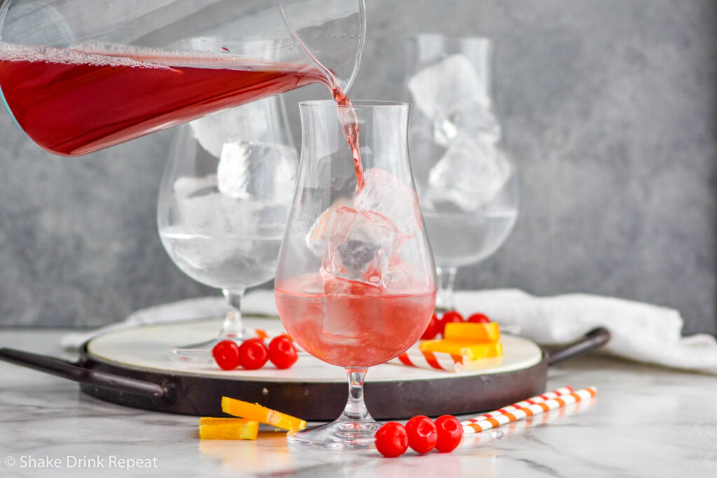 pitcher of cranberry juice pouring into a glass of ice and vodka to make a Sex on The Beach cocktail surrounded by straws, maraschino cherries and orange slices