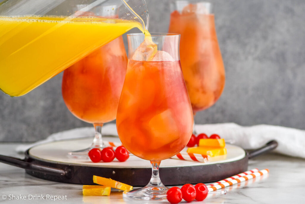 pitcher of orange juice pouring into a glass of Sex on The Beach cocktail surrounded by two more cocktails, straws, maraschino cherries, and orange slices