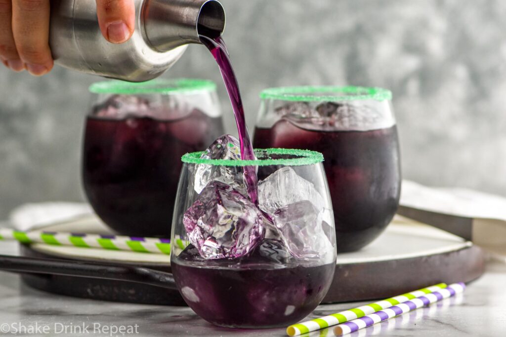 man's hand pouring cocktail shaker of Drunk Witch ingredients into a rimmed glass with ice surrounded by other Drunk Witch cocktails and straws