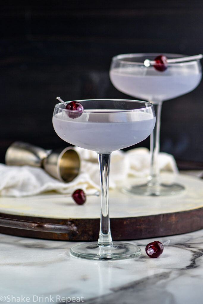 two glasses of Aviation Cocktail garnished with a cherry with cocktail jigger in the background