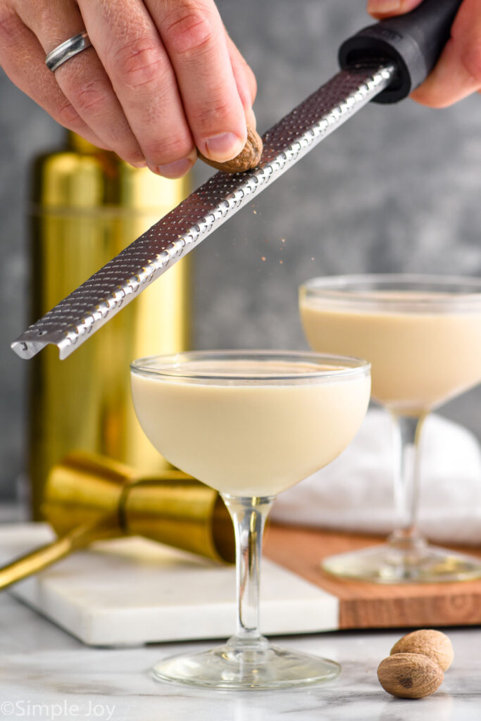 man's hands grating fresh nutmeg into a glass of Brandy Alexander recipe with cocktail shaker and jigger in background