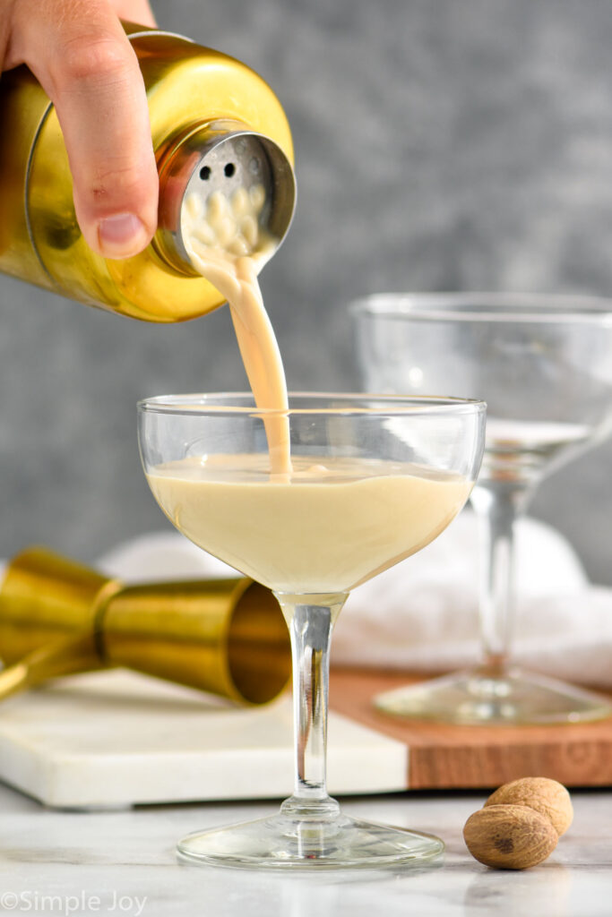 man's hand pouring cocktail shaker of Brandy Alexander ingredients into a glass surrounded by fresh nutmeg