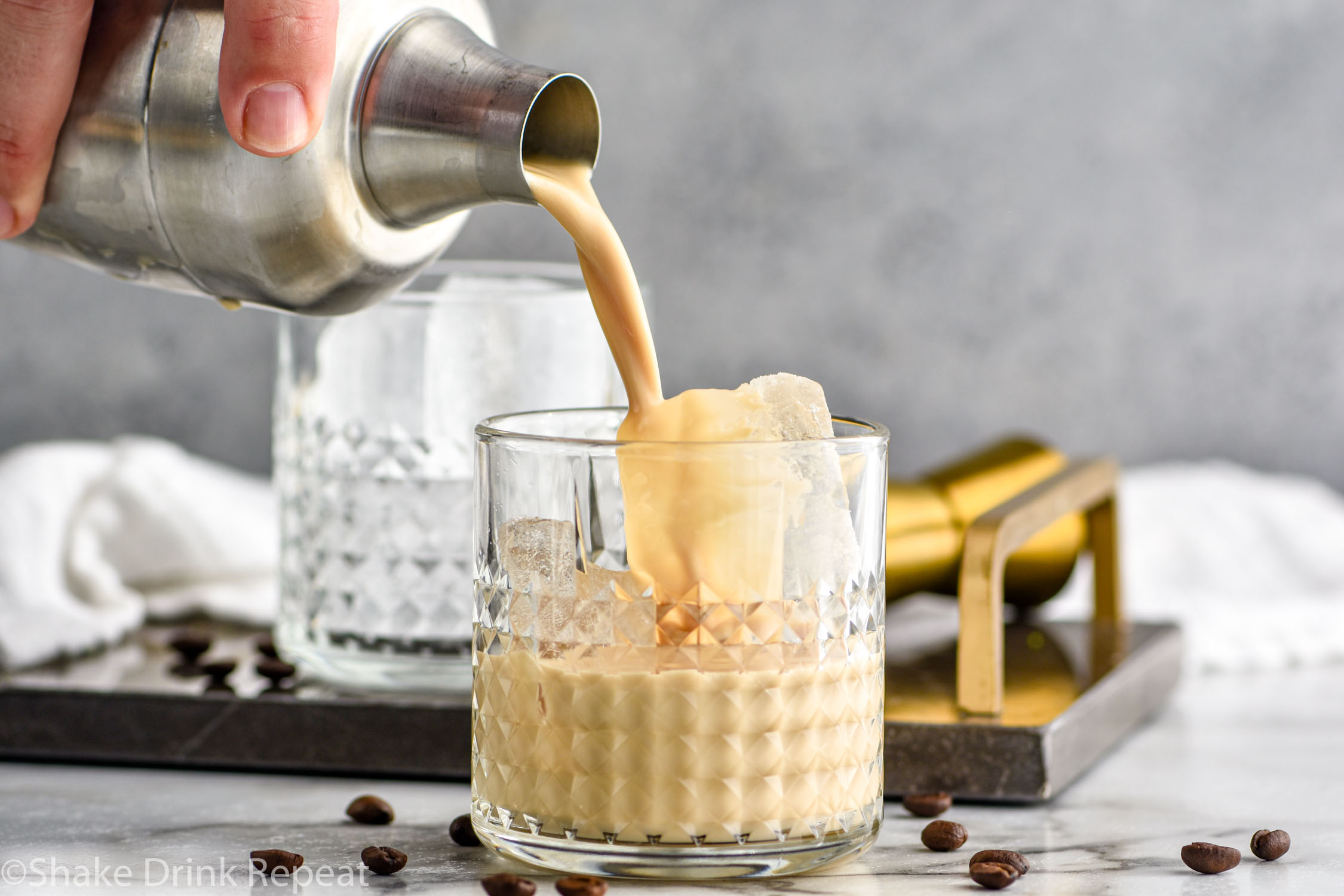 man's hand pouring cocktail shaker of Colorado Bulldog ingredients into a glass of ice surrounded by coffee beans