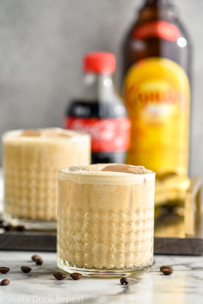 two glasses of Colorado Bulldog recipe with ice surrounded by coffee beans, a bottle of cola, and a bottle of Kahlua