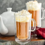 two mugs of Hot Buttered Rum recipe topped with whipped cream and nutmeg and surrounded by cinnamon sticks and a kettle