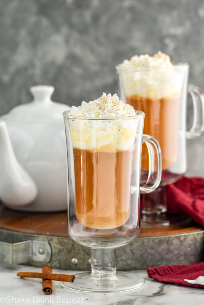 two mugs of Hot Buttered Rum recipe topped with whipped cream and nutmeg and surrounded by cinnamon sticks and a kettle