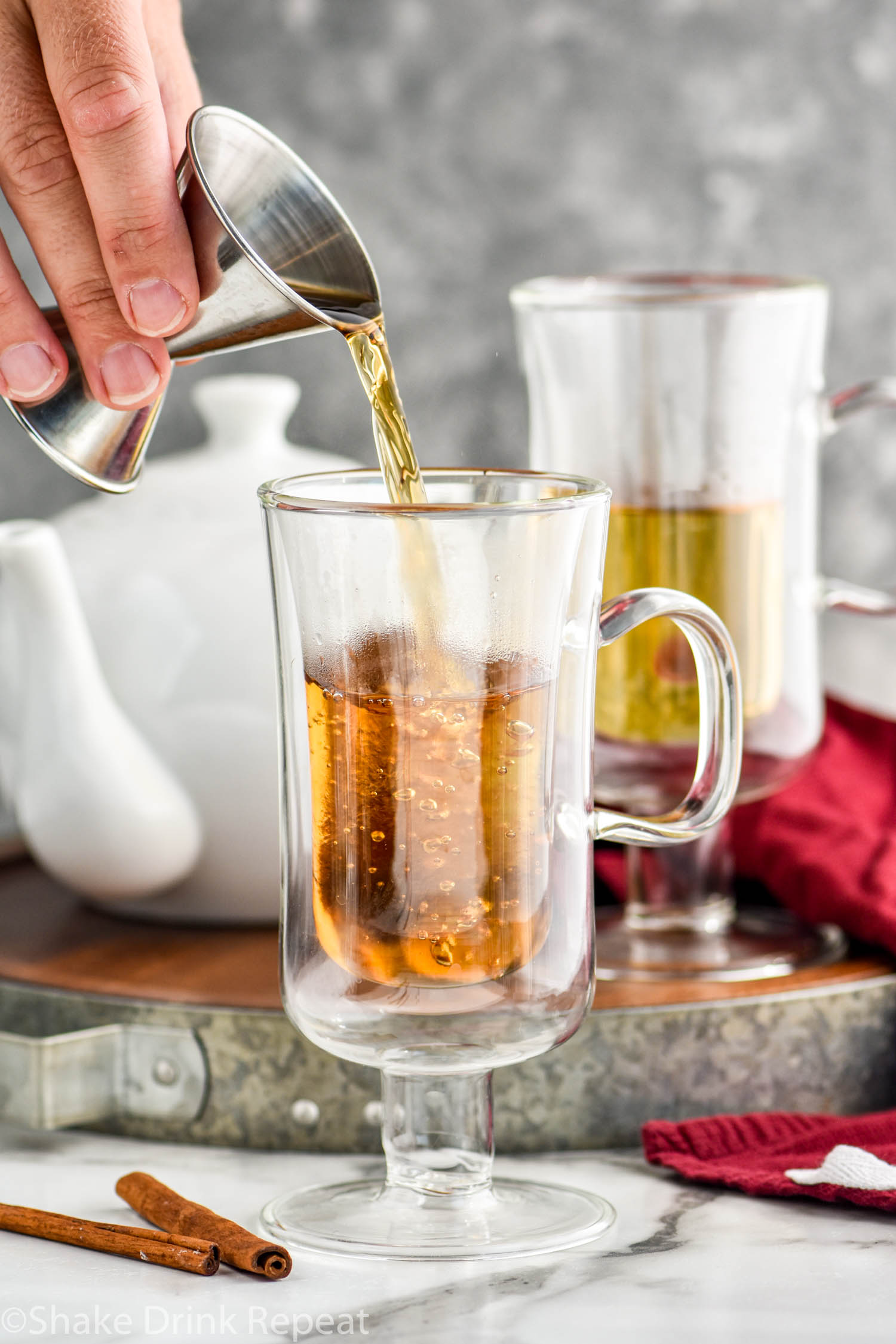 man's hand pouring jigger of rum into a glass of Hot Buttered Rum recipe surrounded by cinnamon sticks and a kettle of hot water
