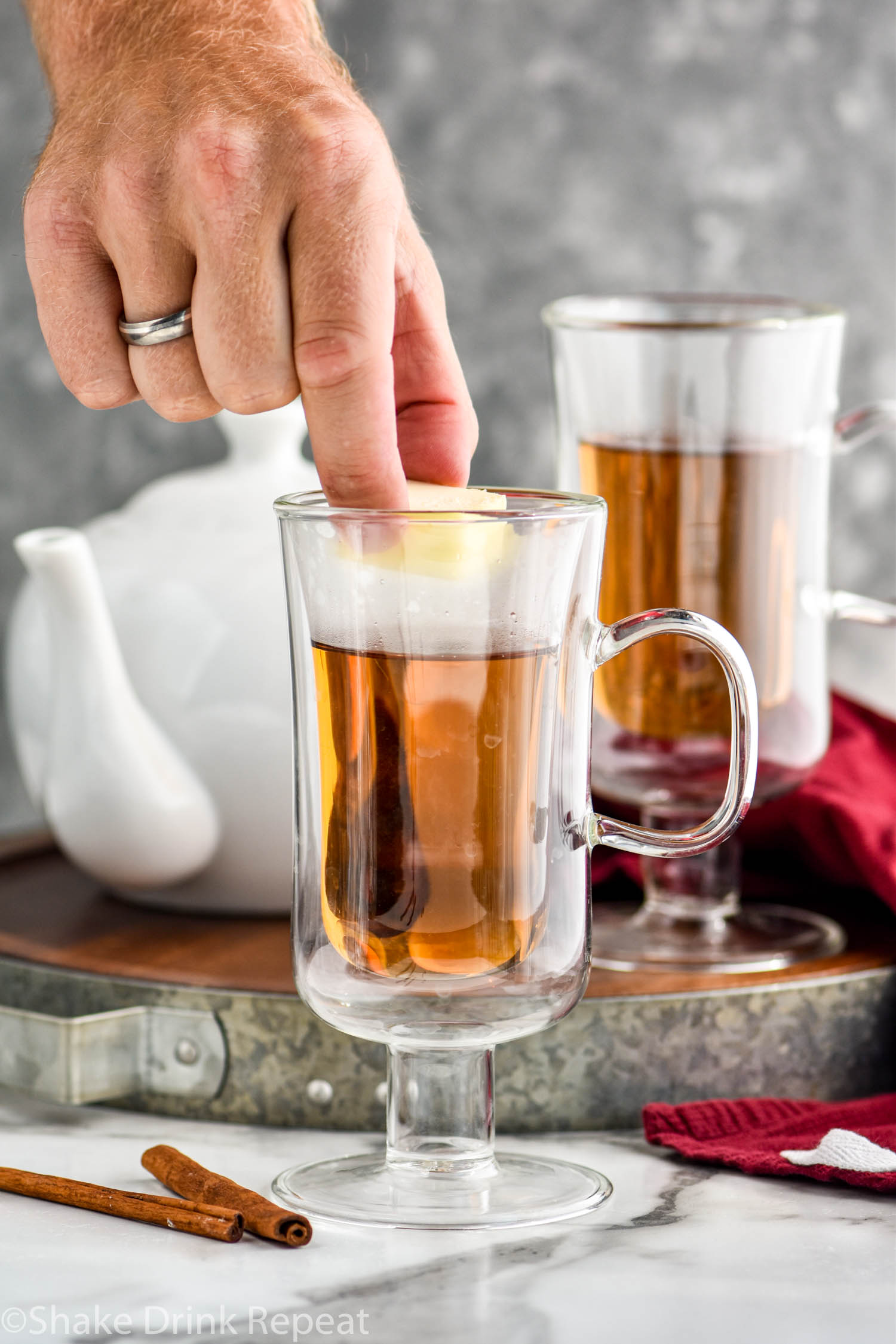 two mugs of Hot Buttered Rum recipe with man's hand placing butter into one of the mugs. Cinnamon sticks and a kettle of hot water in the background