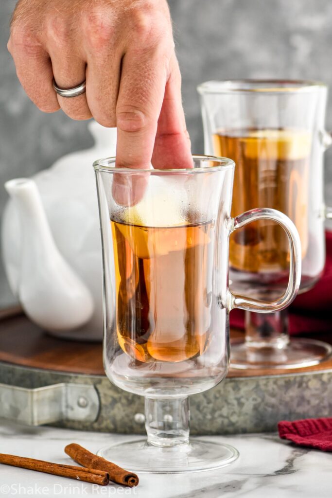 man's hand placing butter into a mug of Hot Buttered Rum ingredients surrounded by cinnamon sticks and a kettle of hot water