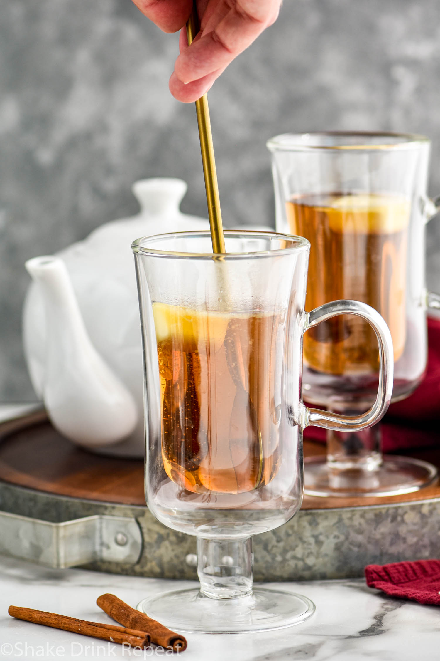 man's hand stirring Hot Buttered Rum recipe in a mug surrounded by cinnamon sticks and a kettle of hot water