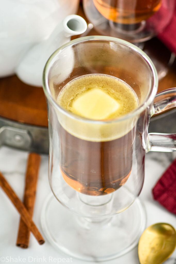 mug of Hot Buttered Rum surrounded by cinnamon sticks and a kettle of hot water