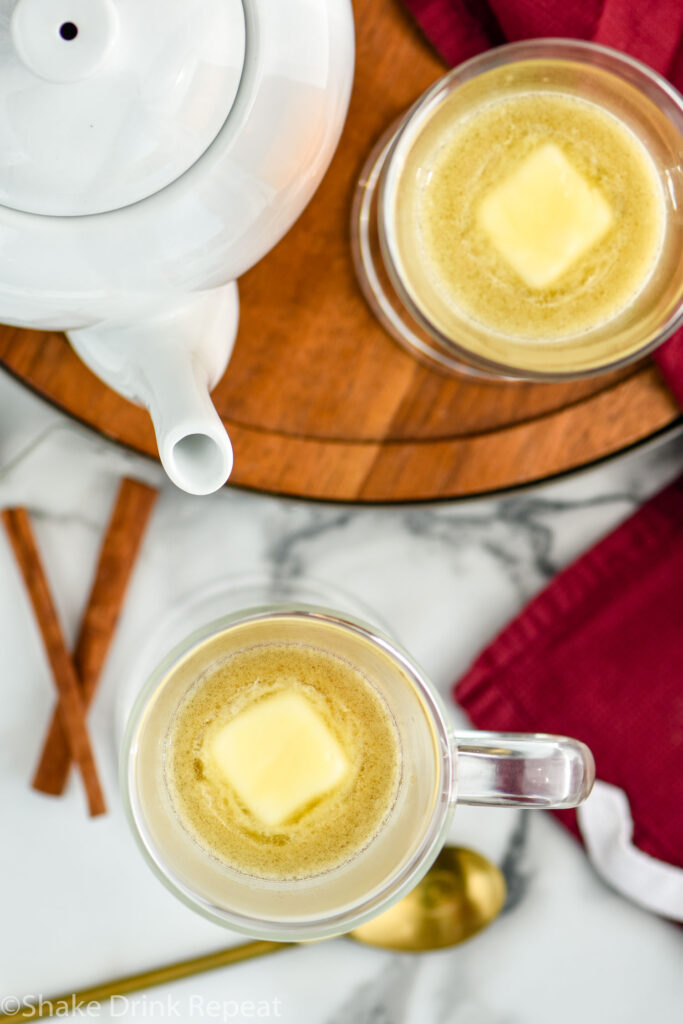 two mugs of Hot Buttered Rum recipe surrounded by cinnamon sticks, a kettle, and a gold spoon