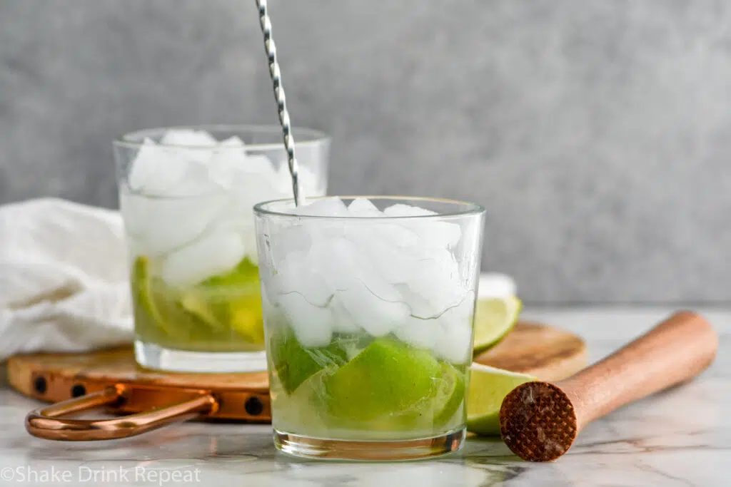 stir stick stirring glass of Caipirinha cocktail recipe with ice and fresh limes