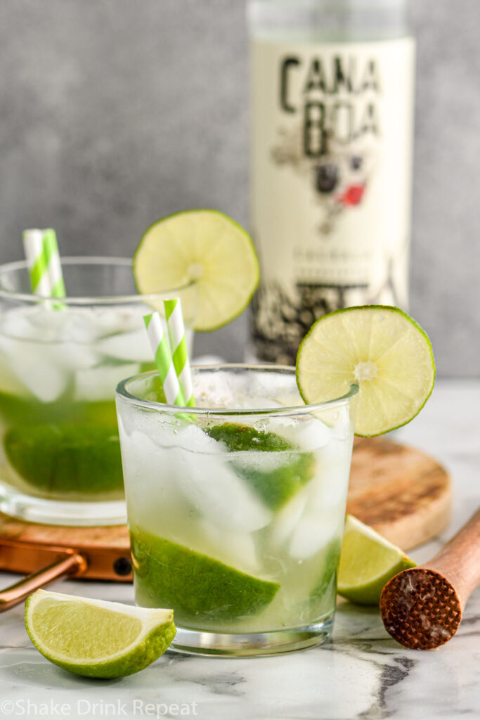 two glasses of Caipirinha with ice, fresh limes, straws, and a bottle of Cachaça in the background