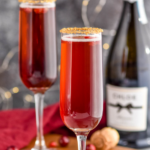 two glasses of Cranberry Pomegranate Champagne Cocktail recipe rimmed with gold sprinkles surrounded by pomegranate seeds and cranberries