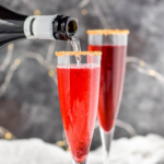 bottle of champagne pouring into a glass of Poinsettia Cocktail recipe with gold sprinkles on the rim surrounded by cranberries