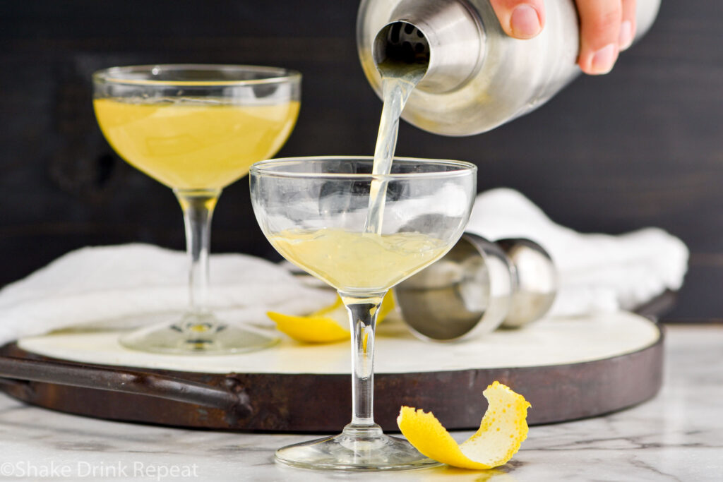 man's hand pouring cocktail shaker of Between the Sheets recipe into a coupe glass surrounded by fresh lemon peel