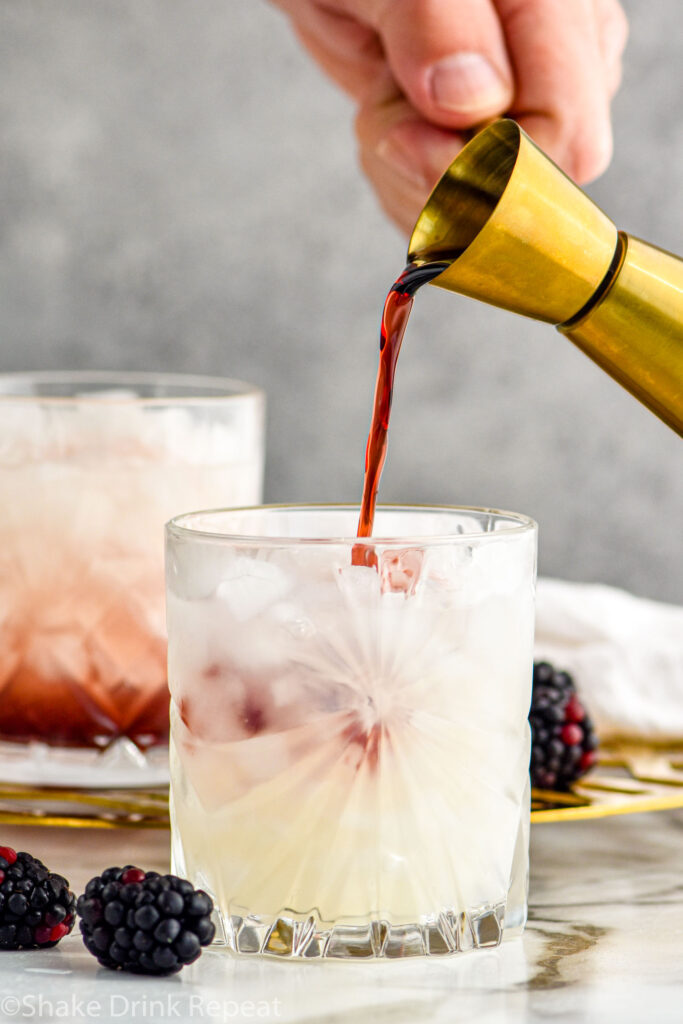 man's hand pouring jigger of Creme de Mure into a glass of Bramble ingredients surrounded by fresh blackberries