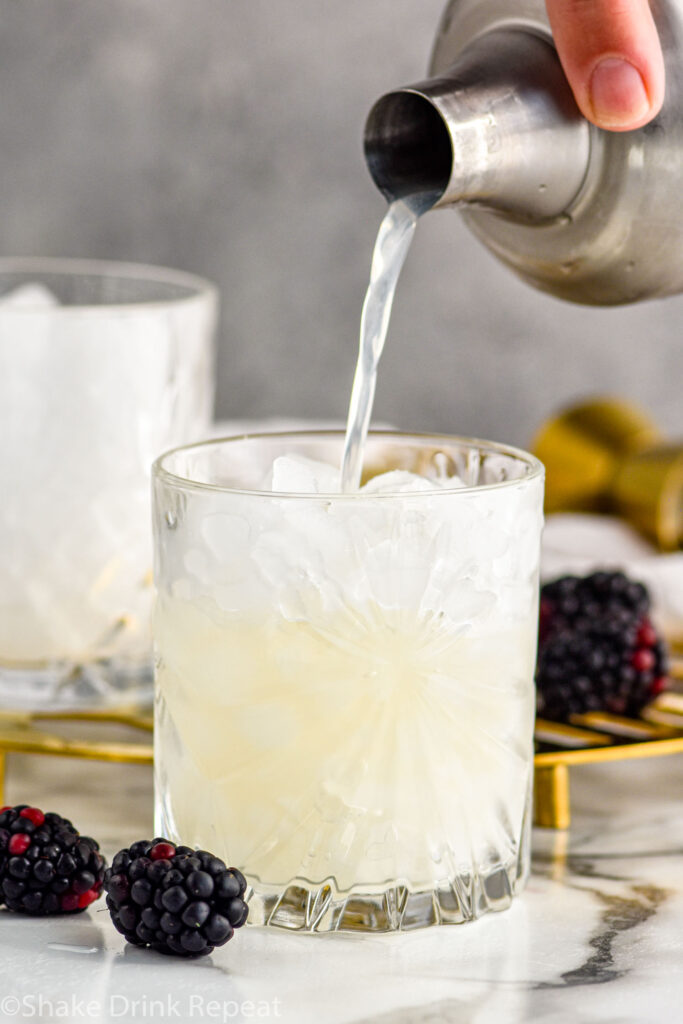 man's hand pouring cocktail shaker of Bramble ingredients into a glass of crushed ice surrounded by fresh blackberries