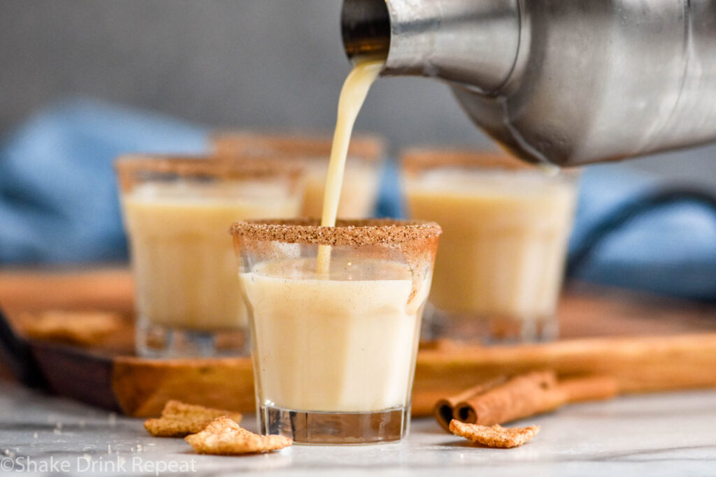 cocktail shaker pouring Cinnamon Toast Crunch Shot ingredients into a shot glass rimmed with cinnamon sugar surrounded by cereal and cinnamon sticks