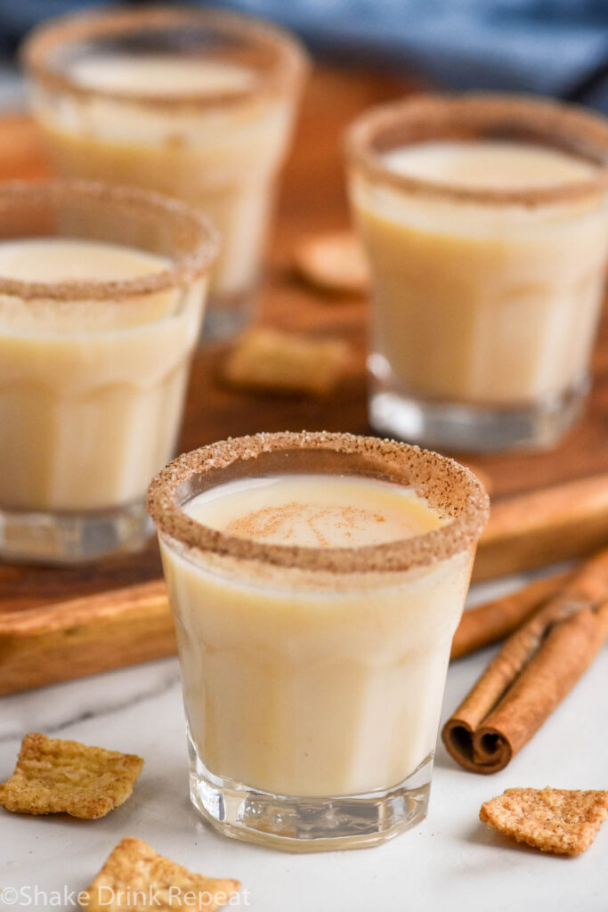 four shot glasses of Cinnamon Toast Crunch Shot recipe with cinnamon sugar rim and whipped cream topping surrounded by cereal and cinnamon sticks