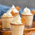 four shot glasses of Cinnamon Toast Crunch Shot recipe with cinnamon sugar rim and whipped cream topping surrounded by cereal and cinnamon sticks