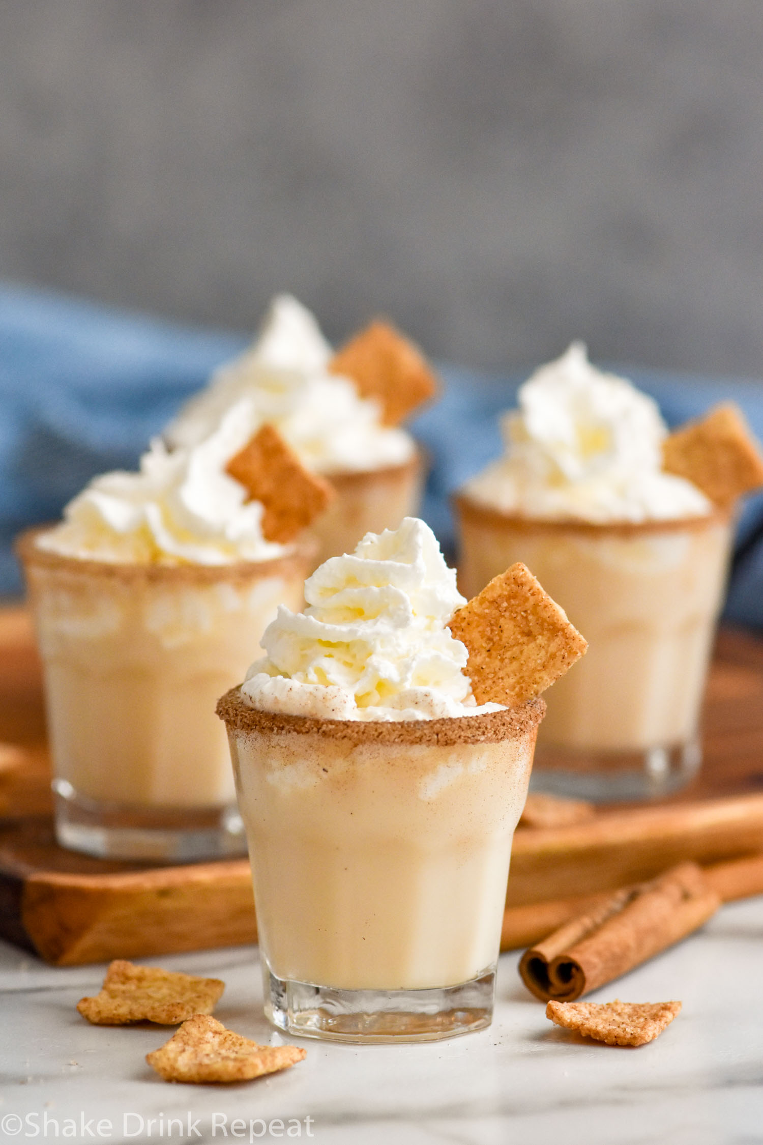 four shot glasses of Cinnamon Toast Crunch Shot recipe with cinnamon sugar rim and whipped cream topping surrounded by cereal and cinnamon sticks