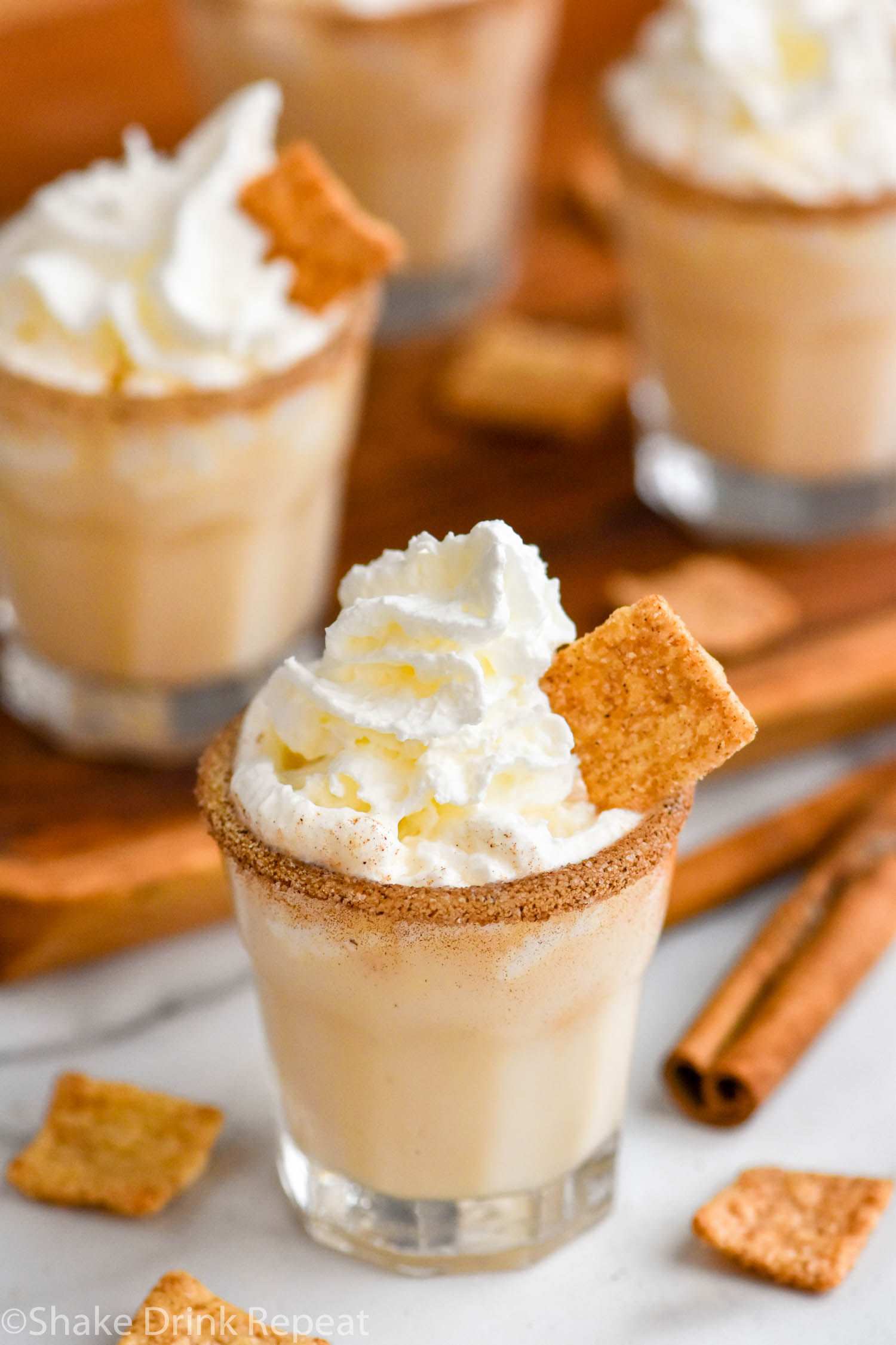 four shot glasses of Cinnamon Toast Crunch Shot recipe with cinnamon sugar rim and whipped cream topping surrounded by cereal and cinnamon sticks