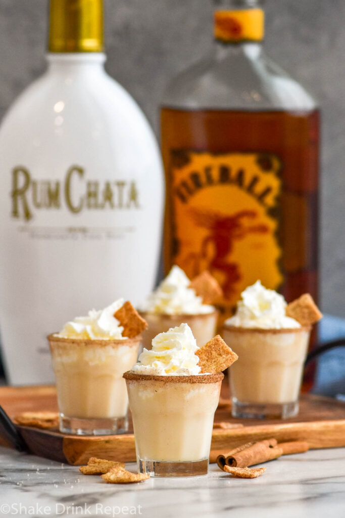 four shot glasses of Cinnamon Toast Crunch Shot recipe with cinnamon sugar rim and whipped cream topping and bottles of RumChata and Fireball whiskey in the background