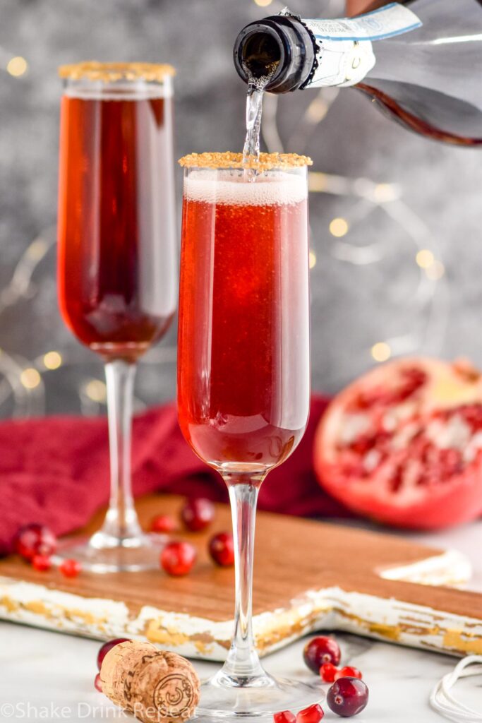 bottle of champagne pouring into a glass of Cranberry Pomegranate Champagne Cocktail recipe rimmed with gold sprinkles surrounded by pomegranate seeds and cranberries