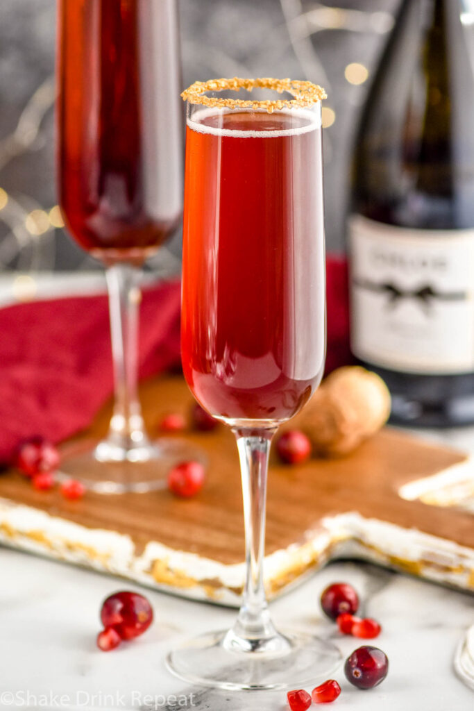 glass of Cranberry Pomegranate Champagne Cocktail recipe rimmed with gold sprinkles surrounded by pomegranate seeds and cranberries