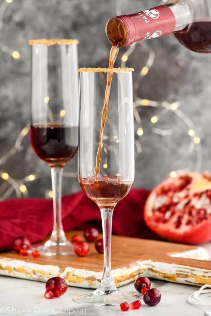 a bottle of pomegranate liqueur pouring into a glass of Cranberry Pomegranate Champagne Cocktail recipe rimmed with gold sprinkles surrounded by pomegranate seeds and cranberries